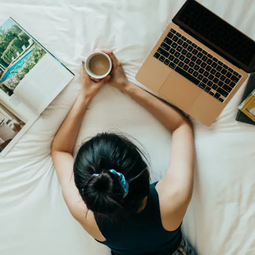 Image of a remote worker  with a book, cup of coffee, and a laptop on a bed